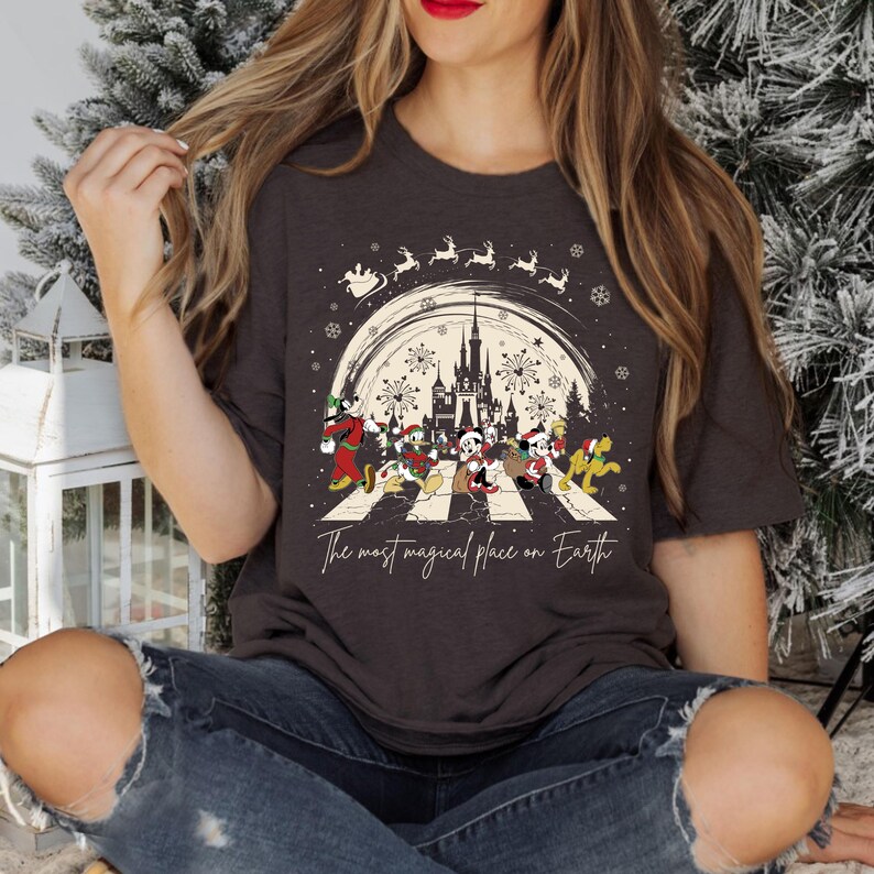 Puede incluir: Una camiseta gris oscuro con un gr&aacute;fico de Mickey Mouse y sus amigos caminando por una calle con el texto "The most magical place on Earth". El gr&aacute;fico tambi&eacute;n incluye una silueta del castillo de Cenicienta y fuegos artificiales.