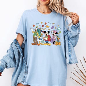 May include: Light blue t-shirt featuring a cartoon illustration of Disney characters Goofy, Donald Duck, Mickey Mouse, Minnie Mouse, and Pluto, all ready for school. The design includes books, leaves, and paper airplanes.