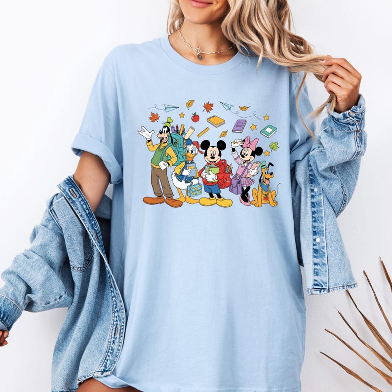 May include: Light blue t-shirt featuring a cartoon illustration of Disney characters Goofy, Donald Duck, Mickey Mouse, Minnie Mouse, and Pluto, all ready for school. The design includes books, leaves, and paper airplanes.