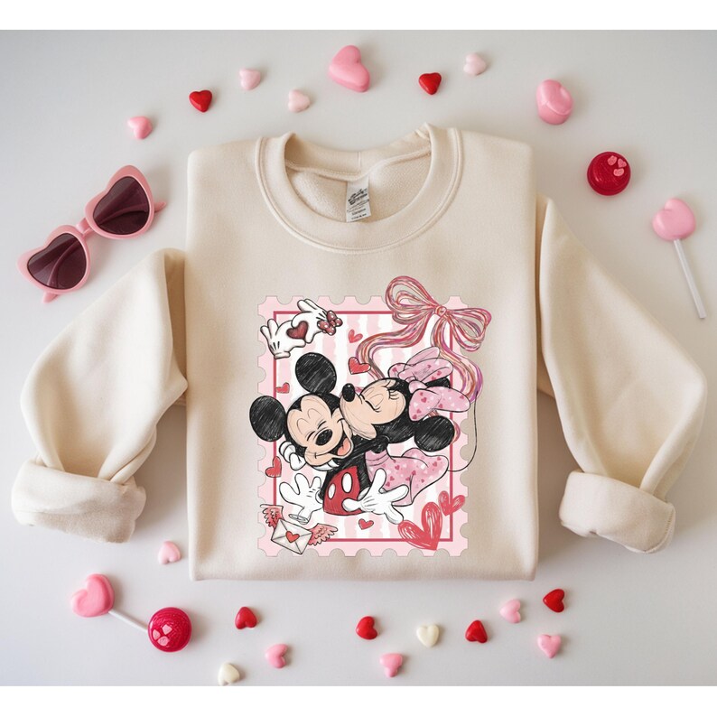 May include: Cream-colored sweatshirt featuring a graphic of Mickey and Minnie Mouse embracing within a pink postage stamp design. The design includes hearts, a bow, and an envelope. Pink heart-shaped sunglasses and candies surround the sweatshirt.