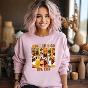 May include: A light pink sweatshirt featuring the text "MICKEY & CO EST. 1928" with a graphic of Disney characters. The graphic includes Donald Duck, Pluto, Mickey Mouse, Minnie Mouse, Goofy, and Donald Duck. The sweatshirt has long sleeves.