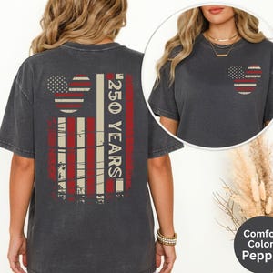 May include: Pepper-colored t-shirt featuring a distressed American flag design with a Mickey Mouse head and the text "250 YEARS" in red and white. The shirt has a crew neck and short sleeves. The shirt is part of the Comfort Colors collection.