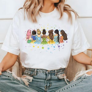 May include: White t-shirt with a colorful illustration of seven princesses standing back to back with stars in the background.