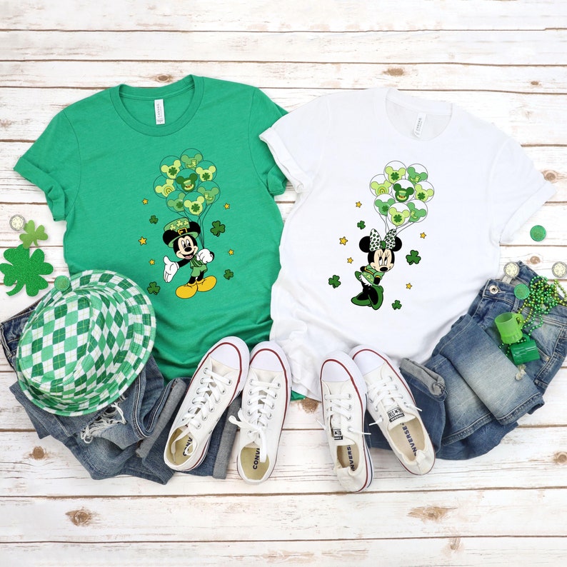 May include: Two green and white t-shirts with a Mickey Mouse and Minnie Mouse design. Mickey Mouse is wearing a green hat and holding green shamrock balloons. Minnie Mouse is wearing a green bow and holding green shamrock balloons. The shirts are on a white wooden background with jeans, a hat, and sneakers.