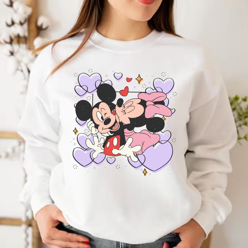 May include: White sweatshirt with a graphic print of Mickey Mouse and Minnie Mouse kissing. The graphic is surrounded by purple hearts and sparkles.
