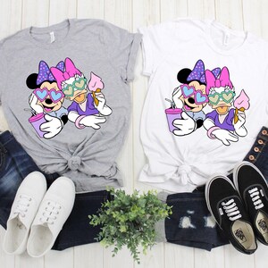 Pode incluir: Duas camisetas cinzas e brancas com uma estampa gr&aacute;fica de Minnie Mouse e Daisy Duck usando &oacute;culos de sol em forma de cora&ccedil;&atilde;o. As camisetas est&atilde;o dispostas sobre uma superf&iacute;cie de madeira com jeans azul e t&ecirc;nis brancos.