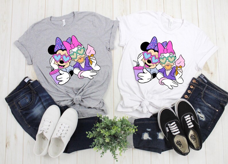 Pode incluir: Duas camisetas cinzas e brancas com uma estampa gr&aacute;fica de Minnie Mouse e Daisy Duck usando &oacute;culos de sol em forma de cora&ccedil;&atilde;o. As camisetas est&atilde;o dispostas sobre uma superf&iacute;cie de madeira com jeans azul e t&ecirc;nis brancos.
