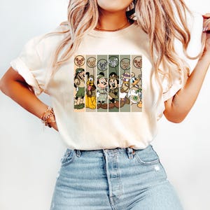 May include: A white t-shirt with a vintage-style graphic of Mickey Mouse, Minnie Mouse, Donald Duck, Goofy, and Pluto dressed in safari outfits. The characters are standing in front of a striped background with animal print balloons.