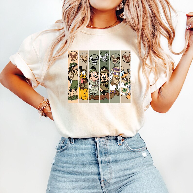 May include: A white t-shirt with a vintage-style graphic of Mickey Mouse, Minnie Mouse, Donald Duck, Goofy, and Pluto dressed in safari outfits. The characters are standing in front of a striped background with animal print balloons.