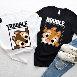 May include: Two t-shirts with a cartoon chipmunk peeking out of a frame. The white shirt says "Trouble" and the black shirt says "Double".