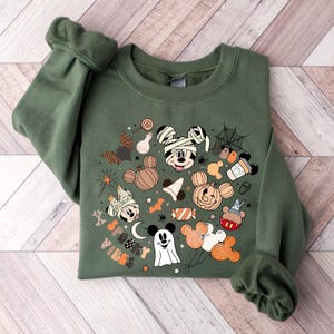 May include: A green sweatshirt with a Halloween-themed Mickey Mouse design. The design features Mickey Mouse in various costumes, including a mummy, a ghost, and a pumpkin. The text "Spooky Vibes" is also included in the design.