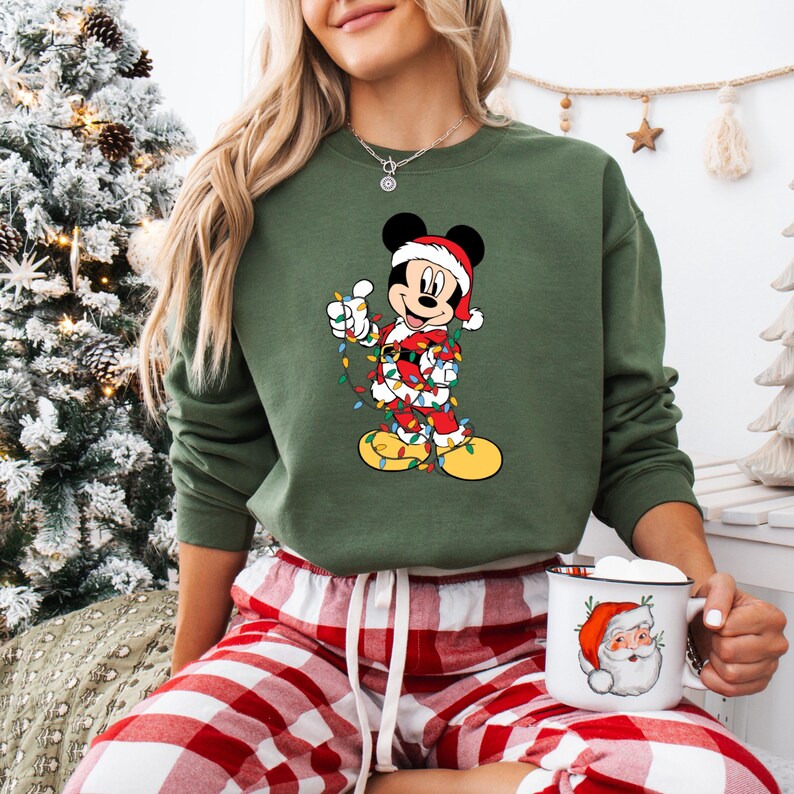May include: Green sweatshirt featuring a festive Mickey Mouse design wrapped in Christmas lights and wearing a Santa hat. The image also shows a mug with a Santa Claus illustration and red and white plaid pajama pants.