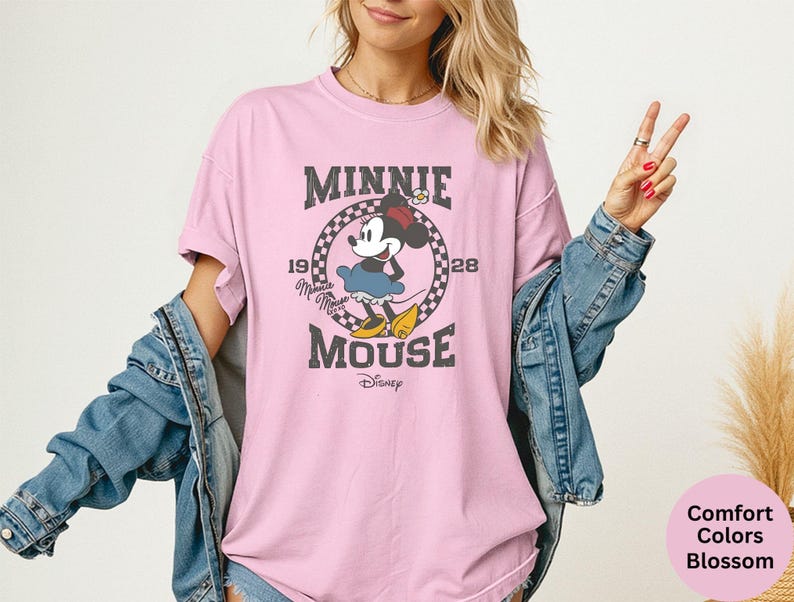 May include: A light pink t-shirt featuring a distressed graphic of Minnie Mouse. The graphic includes the text "MINNIE MOUSE" and the year "1928" in a circular design. The shirt is a Comfort Colors Blossom.