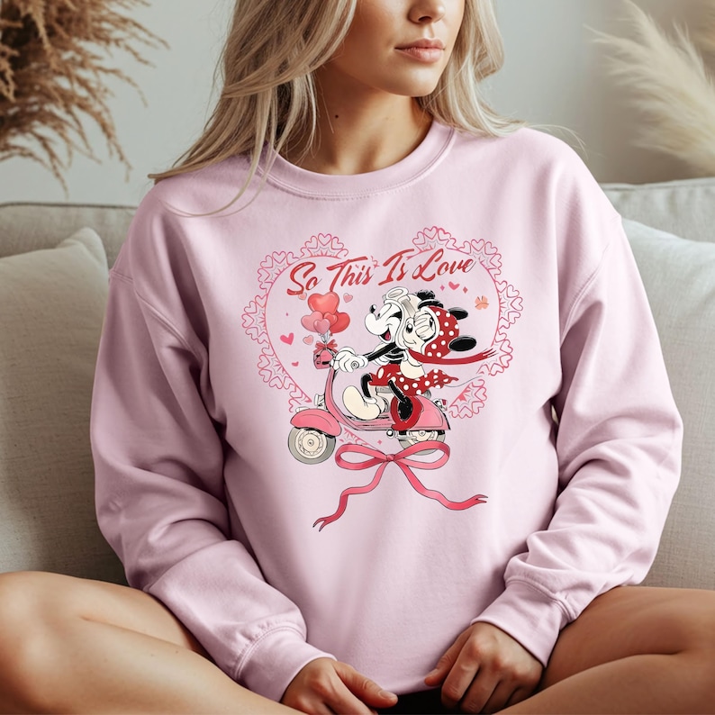 Puede incluir: Sudadera rosa claro con una ilustraci&oacute;n de dibujos animados de una pareja en una moto rosa. El dise&ntilde;o incluye el texto "So This Is Love" dentro de un coraz&oacute;n, con corazones y un lazo.