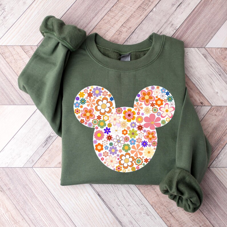 May include: An olive green sweatshirt featuring a floral Mickey Mouse design. The design is composed of various colorful flowers in shades of orange, pink, purple, and yellow. The sweatshirt has long sleeves with ribbed cuffs.