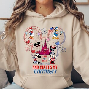 May include: Beige hoodie featuring a colorful graphic of Mickey and Minnie Mouse, Donald and Daisy Duck, and a castle, with the text "4th of July AND YES IT'S MY BIRTHDAY". The design incorporates red, white, and blue elements.