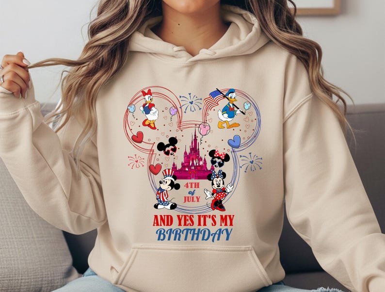 May include: Beige hoodie featuring a colorful graphic of Mickey and Minnie Mouse, Donald and Daisy Duck, and a castle, with the text "4th of July AND YES IT'S MY BIRTHDAY". The design incorporates red, white, and blue elements.