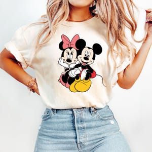 May include: A white t-shirt with a graphic of Mickey Mouse and Minnie Mouse holding hands. The graphic is in black, red, and yellow.