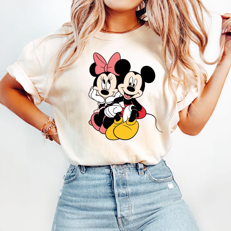 May include: A white t-shirt with a graphic of Mickey Mouse and Minnie Mouse holding hands. The graphic is in black, red, and yellow.