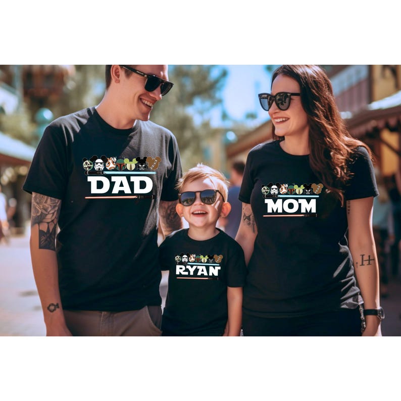 May include: Three people wearing matching black t-shirts with character designs. The shirts read "DAD", "MOM", and "RYAN" in white text. Each shirt has a row of cartoon character faces above the name, with a colorful stripe below.