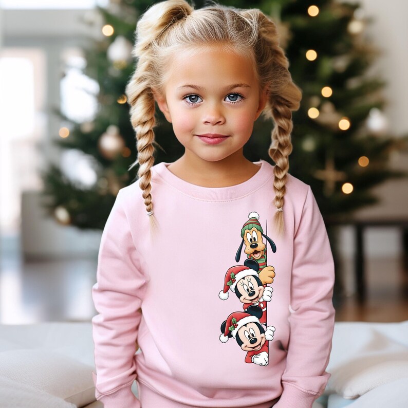 May include: A light pink sweatshirt featuring a graphic of Mickey Mouse, Minnie Mouse, and Pluto wearing Santa hats. The characters are stacked vertically. The background includes a Christmas tree.