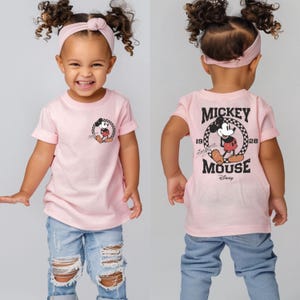 May include: Pink short-sleeved t-shirt with a black and white Mickey Mouse graphic on the front and a black and white Mickey Mouse graphic with the text "Mickey Mouse" on the back.