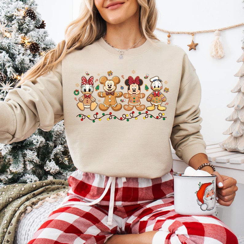 May include: Beige sweatshirt featuring a festive design of Disney characters as gingerbread cookies. The cookies are arranged in a row, with colorful lights below. The sweatshirt is paired with red and white plaid pajama pants.