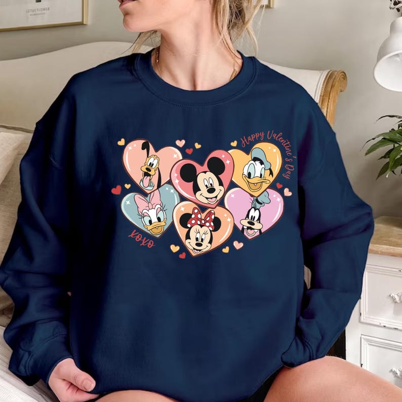 Puede incluir: Una sudadera azul marino con un dise&ntilde;o de San Valent&iacute;n con Mickey Mouse, Minnie Mouse, Donald Duck, Goofy y Pluto. Los personajes est&aacute;n dispuestos en forma de coraz&oacute;n con el texto "Happy Valentine's Day" y "XOXO".
