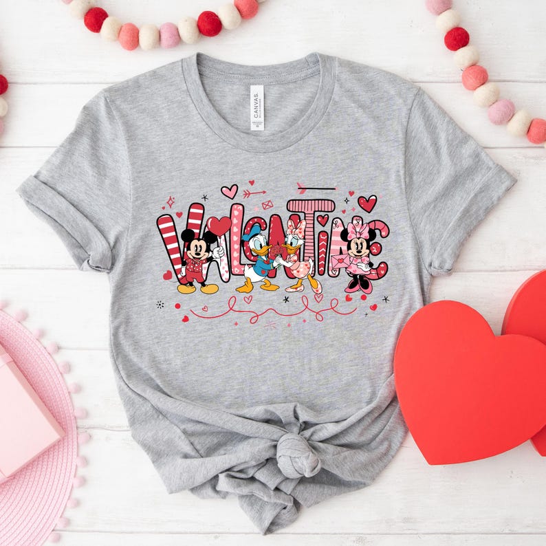 K&ouml;nnte beinhalten: Ein graues T-Shirt mit einem Valentinstags-Design mit Mickey Mouse, Minnie Mouse, Donald Duck und Daisy Duck. Das Design enth&auml;lt das Wort "Valentine" mit Herzen und anderen dekorativen Elementen.