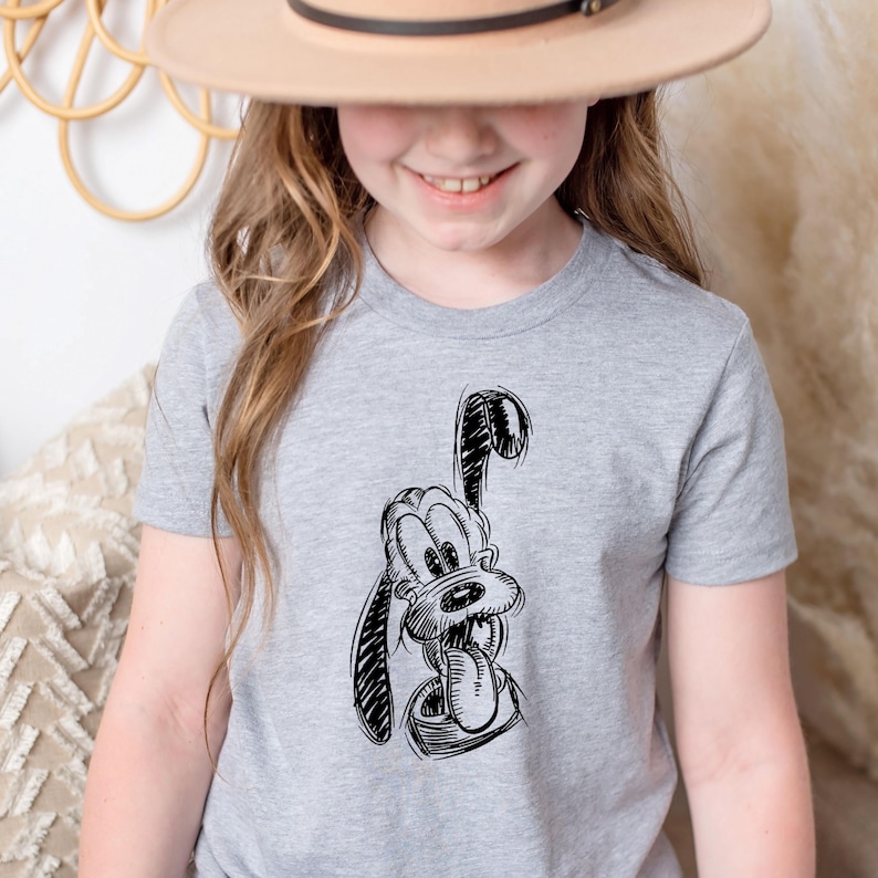 May include: A light gray t-shirt featuring a black and white sketch of a cartoon dog with its tongue sticking out. The dog has large ears and a friendly expression. The shirt is a crew neck style.