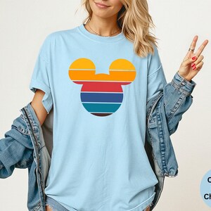 K&ouml;nnte beinhalten: Hellblaues T-Shirt mit einem Retro-Mickey-Mouse-Kopf-Design. Das Design hat horizontale Streifen in Orange, Gelb, Rot, Blau und Braun. Das Shirt ist ein Comfort Colors Chambray.