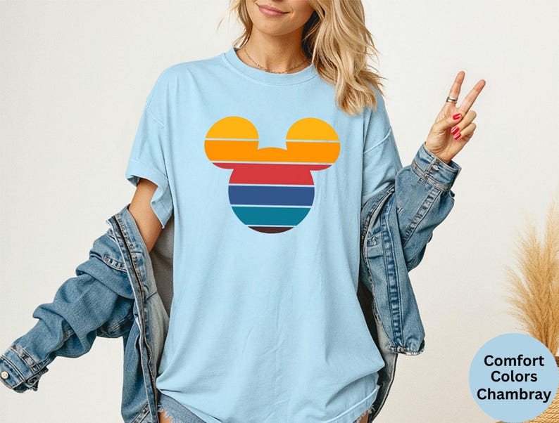 K&ouml;nnte beinhalten: Hellblaues T-Shirt mit einem Retro-Mickey-Mouse-Kopf-Design. Das Design hat horizontale Streifen in Orange, Gelb, Rot, Blau und Braun. Das Shirt ist ein Comfort Colors Chambray.
