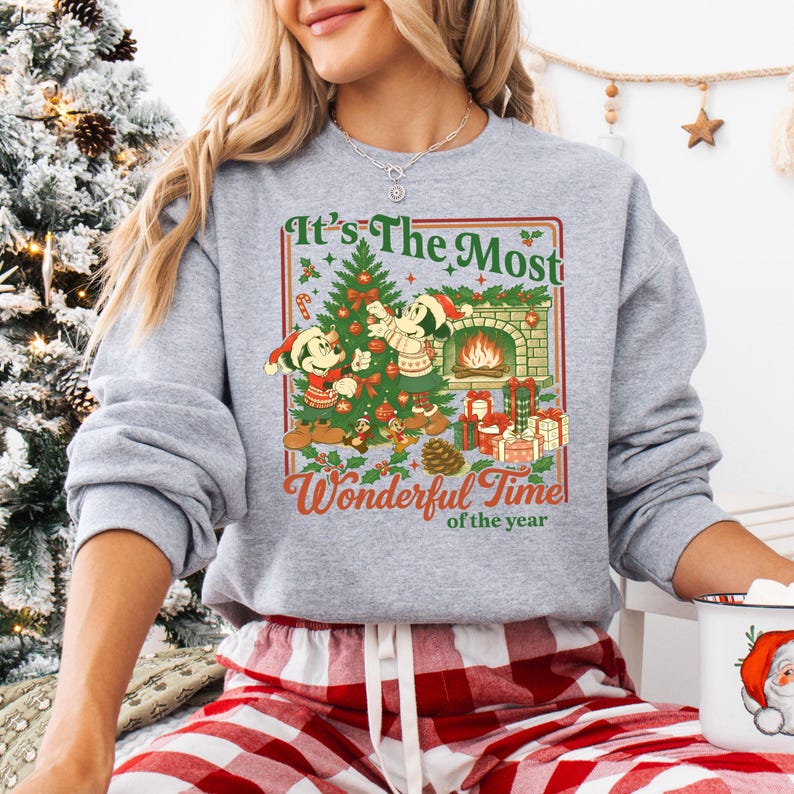 May include: A grey sweatshirt with a Christmas graphic. The design includes a Christmas tree, a fireplace, and the text "It's The Most Wonderful Time of the Year." The graphic features Mickey and Minnie Mouse characters in festive outfits.