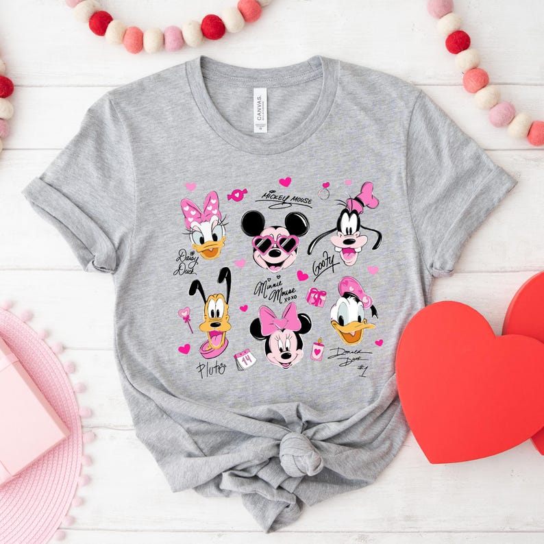 May include: A gray t-shirt featuring a collection of Disney characters, including Mickey Mouse, Minnie Mouse, Donald Duck, and Goofy. The design incorporates pink hearts and character signatures. The shirt is tied in a knot at the bottom.