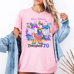 May include: Pink t-shirt with a colorful graphic of a cartoon duck, Disneyland text, and the words "Walt Disney" and "Disneyland 70".