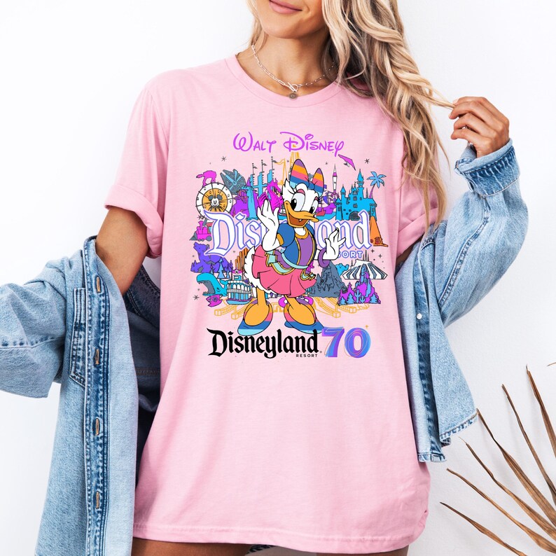 May include: Pink t-shirt with a colorful graphic of a cartoon duck, Disneyland text, and the words "Walt Disney" and "Disneyland 70".