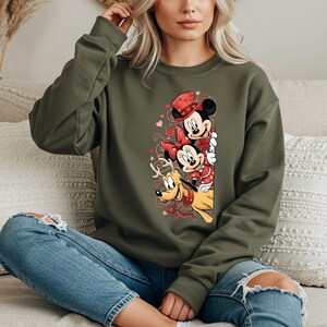 Puede incluir: Una sudadera verde oliva con un dise&ntilde;o de Mickey Mouse, Minnie Mouse y Pluto con atuendos de San Valent&iacute;n. Mickey lleva un sombrero de copa rojo, Minnie un lazo rojo y Pluto un collar rojo. El dise&ntilde;o incluye corazones.