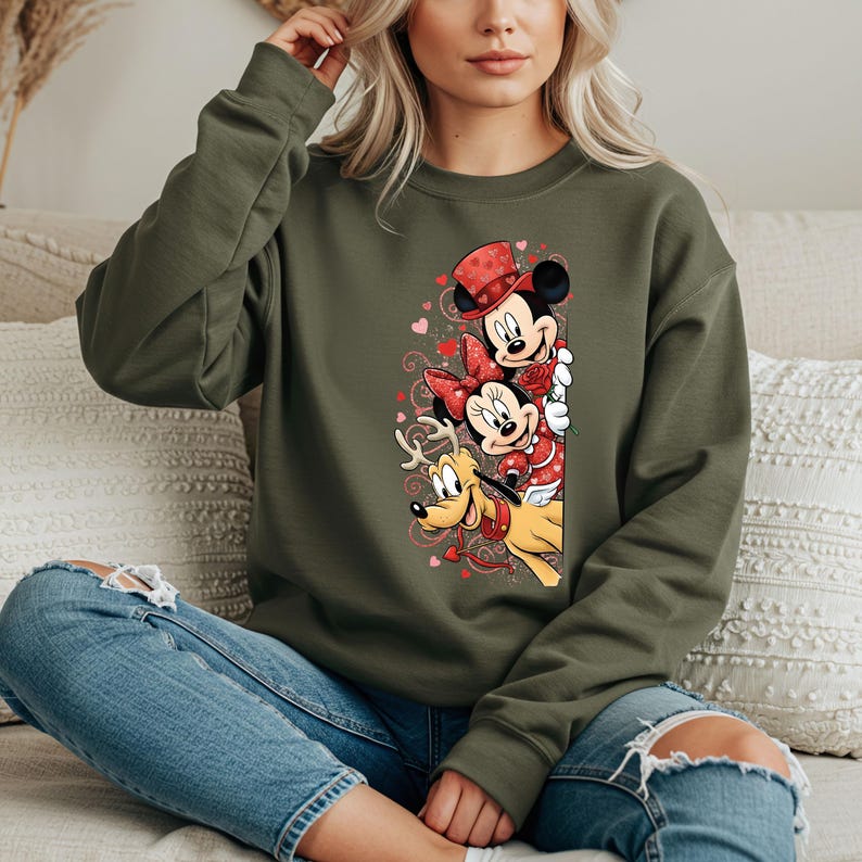 Puede incluir: Una sudadera verde oliva con un dise&ntilde;o de Mickey Mouse, Minnie Mouse y Pluto con atuendos de San Valent&iacute;n. Mickey lleva un sombrero de copa rojo, Minnie un lazo rojo y Pluto un collar rojo. El dise&ntilde;o incluye corazones.