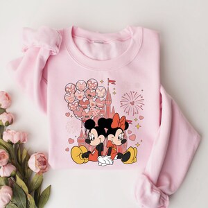 May include: Pink sweatshirt with a graphic of Mickey Mouse and Minnie Mouse in front of a pink castle with hearts and fireworks. The text "LOVE" is on one of the hearts.