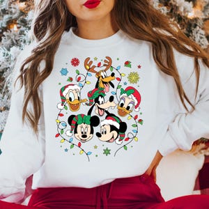 May include: White Christmas sweater with a festive design featuring Mickey Mouse, Minnie Mouse, Donald Duck, Goofy, and Pluto. The characters are surrounded by colorful Christmas lights and snowflakes.