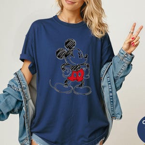 May include: Navy blue t-shirt featuring a sketched Mickey Mouse design with black, red, and white accents. The graphic tee has a crew neck and short sleeves. The shirt is part of the Comfort Colors collection.