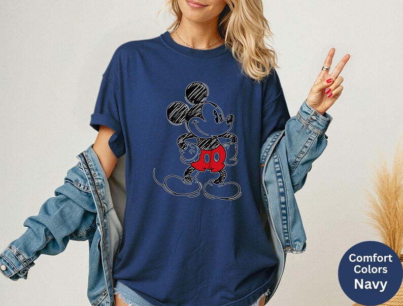 May include: Navy blue t-shirt featuring a sketched Mickey Mouse design with black, red, and white accents. The graphic tee has a crew neck and short sleeves. The shirt is part of the Comfort Colors collection.