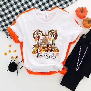 May include: White t-shirt with a colorful Thanksgiving graphic featuring Disney characters, pumpkins, and the words "Happy Thanksgiving" in a script font. The shirt has orange trim and is paired with black pants.