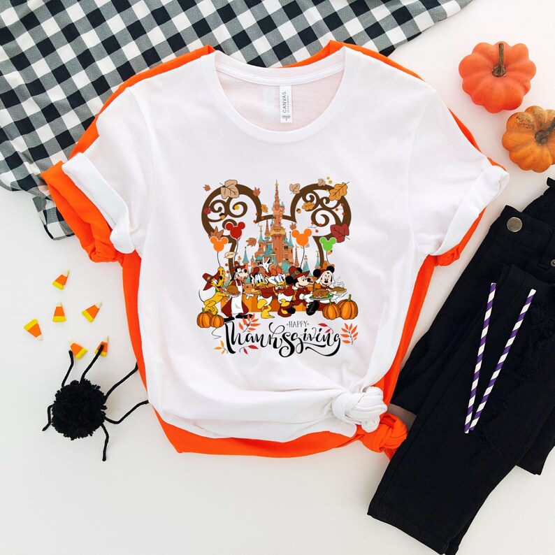 May include: White t-shirt with a colorful Thanksgiving graphic featuring Disney characters, pumpkins, and the words "Happy Thanksgiving" in a script font. The shirt has orange trim and is paired with black pants.