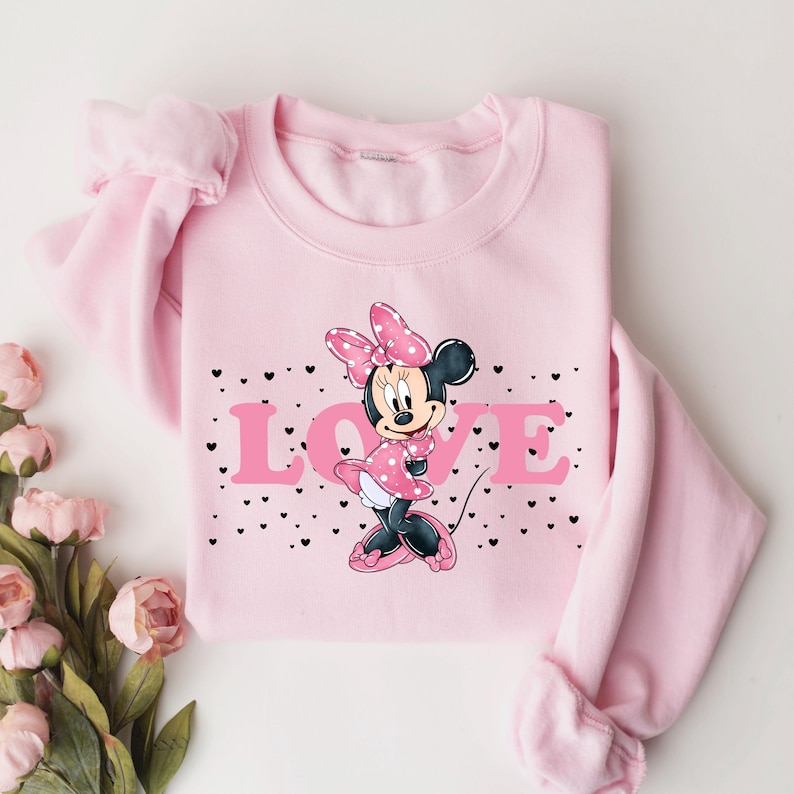 Op de afbeelding: Roze trui met een afbeelding van Minnie Mouse die een roze jurk en een rode strik draagt. De afbeelding bevat ook het woord "LOVE" in roze letters met zwarte contouren. De trui heeft geruite mouwen.