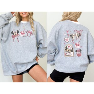 May include: A light gray sweatshirt featuring Minnie Mouse-themed graphics. The front displays Minnie with a bow, hearts, and a bow-shaped headband. The back showcases Minnie and Mickey Mouse with hearts and a cupcake.