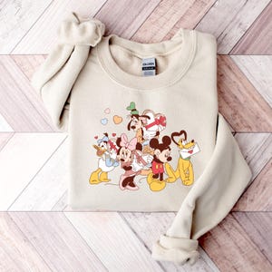 May include: A beige sweatshirt with a graphic print of Mickey Mouse, Minnie Mouse, Donald Duck, Goofy, and Pluto. The characters are holding hearts and gifts. The text "GILDAN" is visible on the tag.