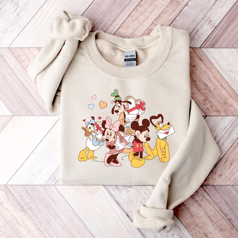 May include: A beige sweatshirt with a graphic print of Mickey Mouse, Minnie Mouse, Donald Duck, Goofy, and Pluto. The characters are holding hearts and gifts. The text "GILDAN" is visible on the tag.