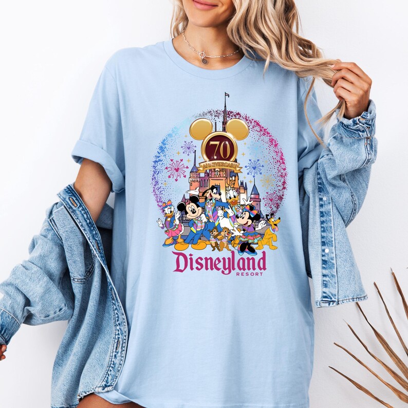 Op de afbeelding: Lichtblauw T-shirt met een afbeelding van Mickey Mouse en zijn vrienden die het 70-jarig jubileum van Disneyland vieren. De afbeelding toont Mickey Mouse, Minnie Mouse, Donald Duck, Goofy, Pluto en andere Disney-figuren voor Sleeping Beauty's Castle. De tekst "Disneyland Resort" is onder de afbeelding gedrukt.