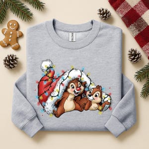 May include: A light gray sweatshirt featuring a festive graphic of two cartoon chipmunks nestled under a Santa hat adorned with colorful Christmas lights. The design is set against a backdrop of holiday decorations.
